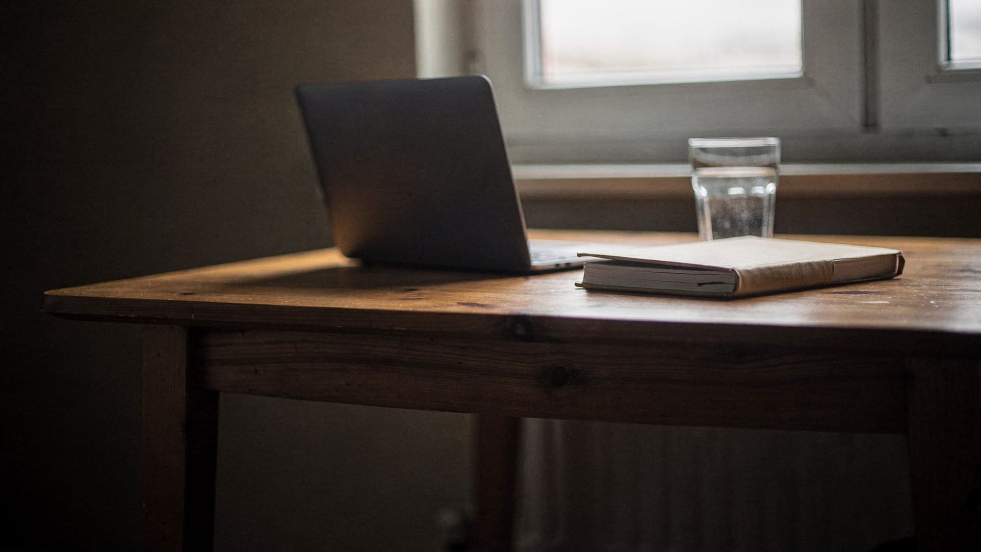 A small wooden table by a window in grey morning light, an open laptop seen from behind, a notebook and a glass of water beside it.