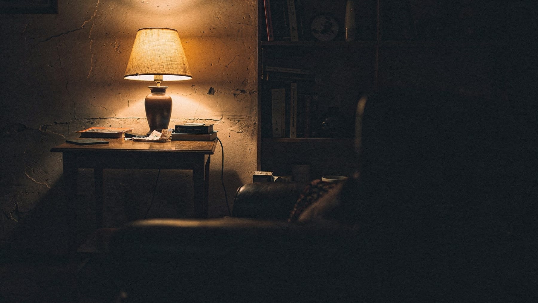 A corner of a room at night lit by a single table lamp, a pool of warm light on a wooden surface, the edge of a bookshelf in shadow.