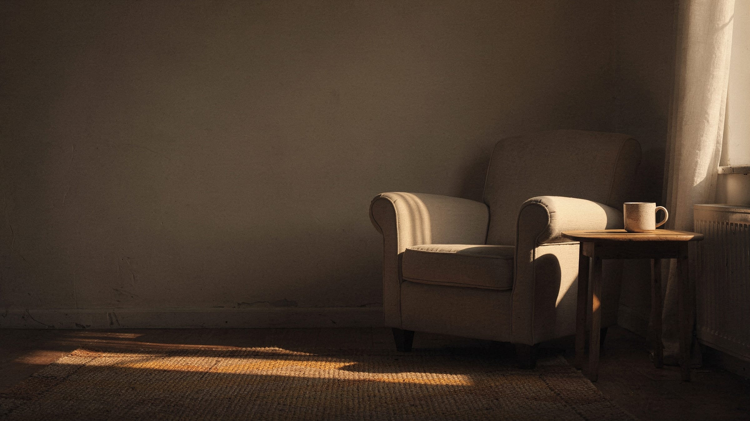The corner of a living room in late afternoon, a comfortable armchair beside a small table with a mug on it, low sun from a window on the right.